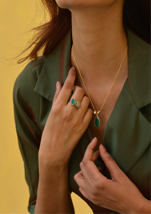Woman wearing green jacket poses elegantly with a gold necklace featuring a green pendant, complemented by a green two-stone ring