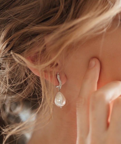 A close-up image of a blonde woman showcasing earrings adorned with white diamond stones. The earrings prominently feature a freshwater pearl pendant. The woman poses from a side angle, with her hands gently touching her face.
