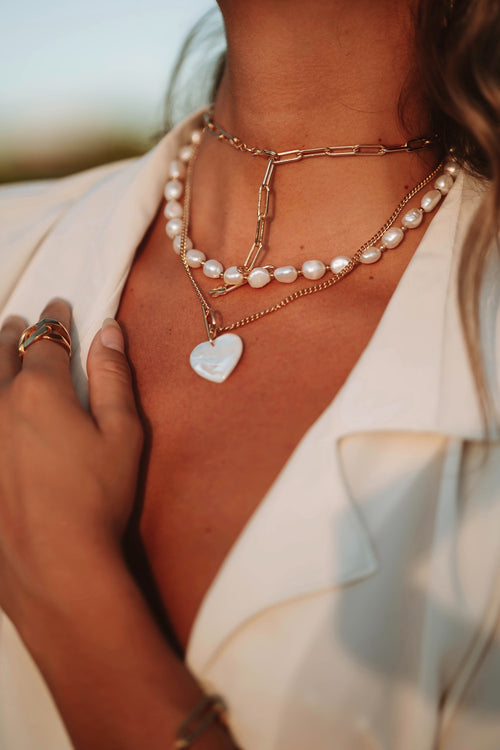 The woman model wearing a white jacket poses elegantly, showcasing a trio of stacking necklaces. The first necklace features a white heart-shaped pendant, followed by a freshwater pearl necklace, and a rectangular gold-colored chain necklace.